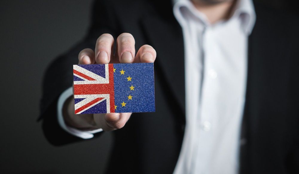Man wearing black open suit jacket and white shirt holding card with half union jack and half EU flag up to the camera. 